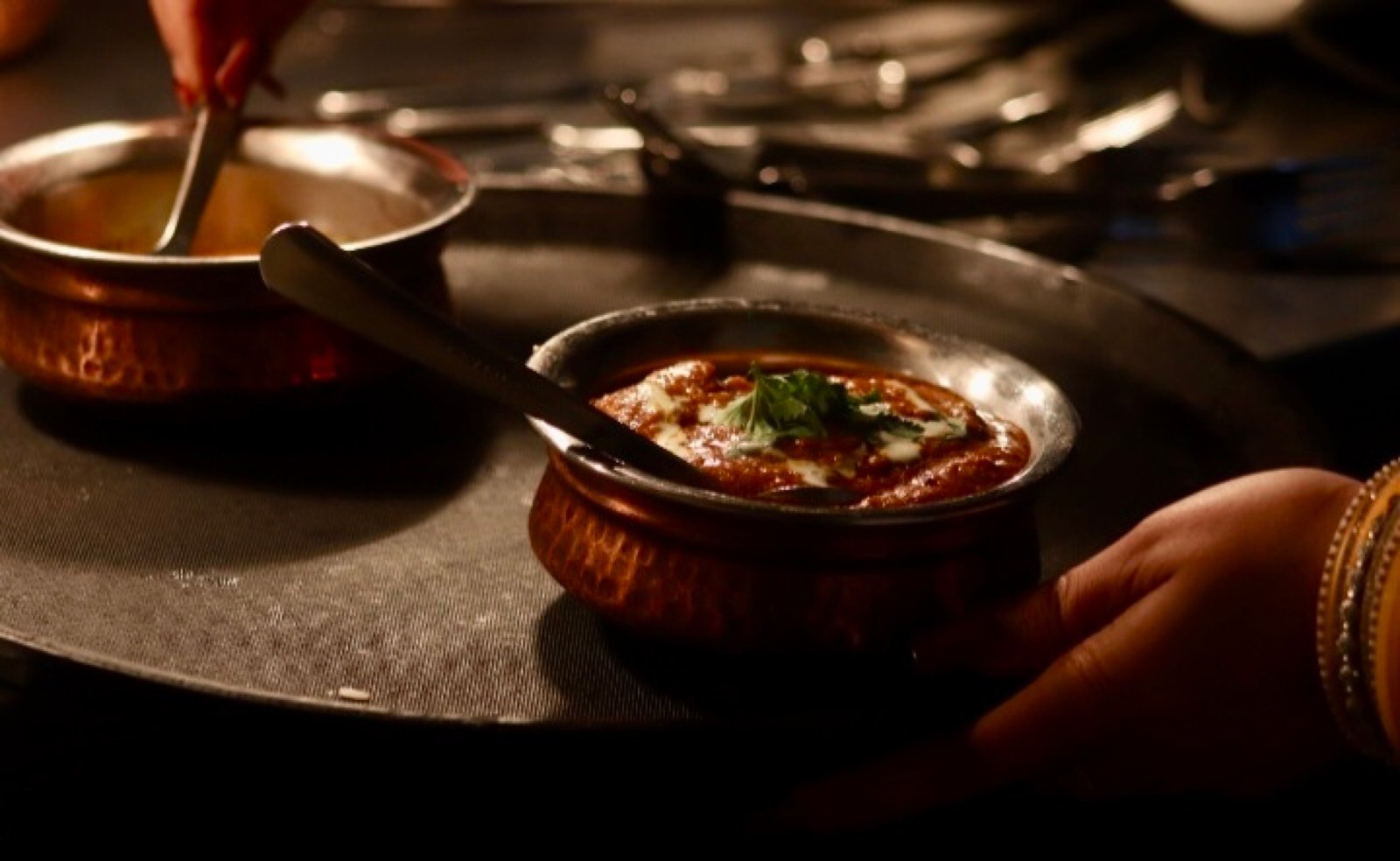A copper handi of curry carried to the table on a dark steel tray, candlelit